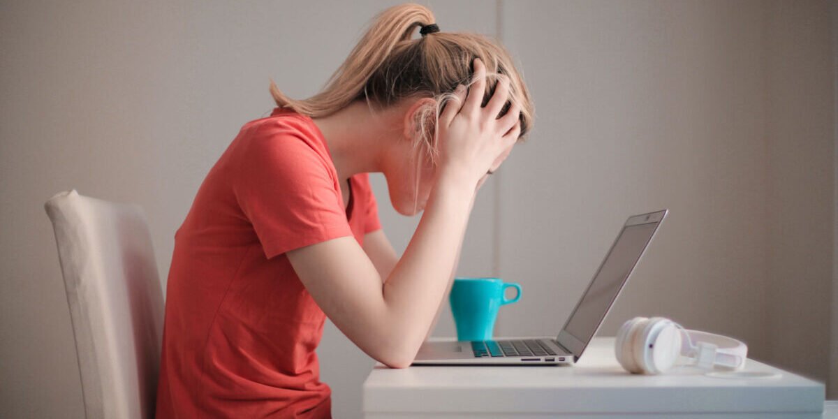 Woman sitting on desk with hands on her head as she looks at a computer