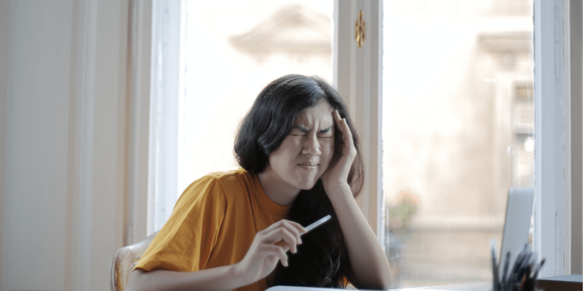 Girl sitting at desk with a pen in hand, closing her eyes and hand to her head